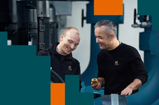 To mænd i arbejdstøj smiler og samarbejder ved industriudstyr i en fabrik.