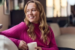 Kvinde med en lyserød trøje, der sidder i en sofa og drikker en kop kaffe.