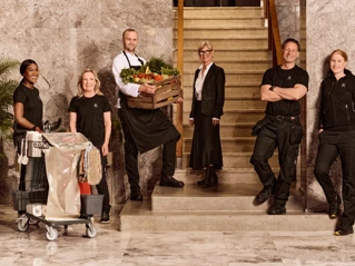 Fem professionelle medarbejdere stiller op i forskellige arbejdspåklædninger foran en marmorvæg og trætrappe, der repræsenterer forskellige brancher, såsom rengøring, gastronomi og byggeri, for at fremvise en mangfoldig arbejdsstyrke.