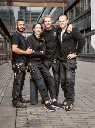 Fire håndværkere i arbejdstøj poserer smilende foran kontorbygning, med værktøjsbælter og firmalogoer.