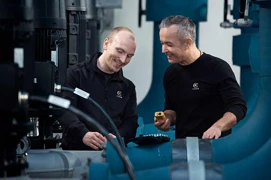 To mænd i arbejdstøj med logo smiler og diskuterer udstyr på en industrivirksomhed.