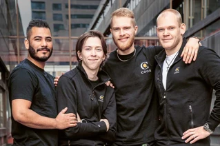 Fire smilende kolleger i sort arbejdstøj med COOR-logo foran et bylandskab.