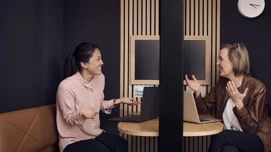 To kvinder smiler og taler sammen på kontoret med bærbare computere.