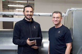 To mænd i arbejdstøj med firma-logo smiler i en industrihal, den ene holder en tablet.
